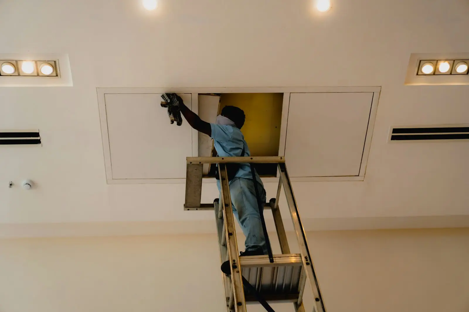 Technician on a ladder opening a ceiling access panel to reach exhaust ductwork